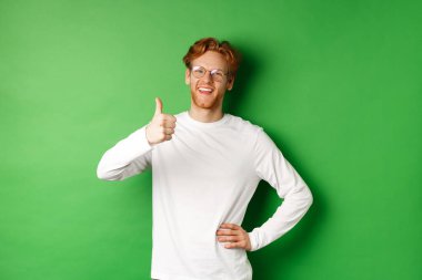 Sakallı, kızıl saçlı, gözlüklü, onaylanmış ve gülümseyen, yeşil arka planda duran neşeli genç adam.