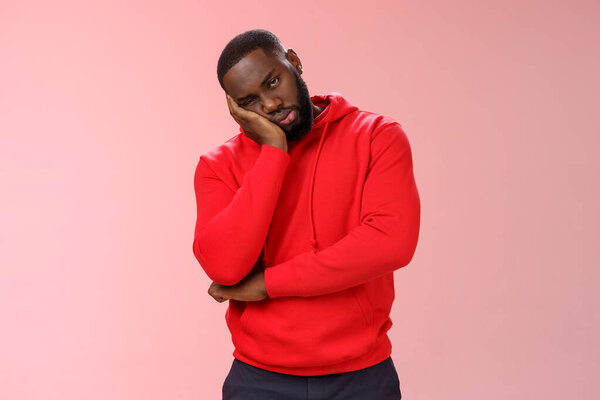 Bored young 25s black guy in red hoodie facepalm lean head hand look indifferent sleepy attend boring meeting fall asleep carelessly listening uninteresting story, pink background