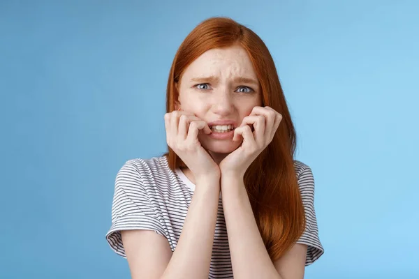 Worried uncomfortable scared young panicking redhead young girl feeling pressure distressed frowning squinting frightened biting fingernails trembling fear, standing blue background