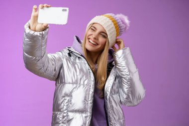 Neşeli, büyüleyici, sarışın, eğlenen kız kayak merkezindeki tatilden resimler yollamak, seflie tutmak, akıllı telefon taklit etmek, göz kırpma, gezici görünüm, mor arka plan.