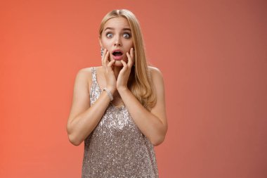 Şok olmuş genç Avrupalı, şaşkın, genç, sarışın kadın gümüş önlüklü, çenesi açık, ağzı açık, ağzı açık, ağzı açık, şaşkın, kafası karışmış, kırmızı arka planda.