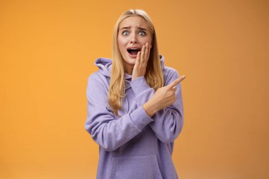 Şaşırtıcı derecede heyecanlı kapüşonlu genç sarışın kız avucunun içi gibi bağırıyor sağa doğru hayret verici bir şekilde haykırıyor ünlü süper star neşeli, pozitif duyguları tutamıyor, turuncu arka plan