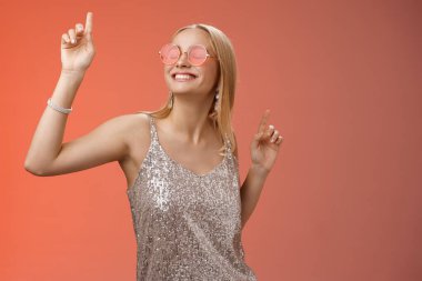 Keyifli, çekici, şık, bin yıllık sarışın, eğlenen, kutlama yapan, güneş gözlüğü takan, gümüş renkli elbiseler giyen, dans eden, geniş bakışlı, el sallayan, kırmızı arka plan.