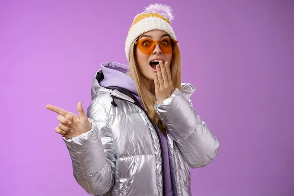 Amazed speechless surprised attractive stylish blond girlfriend in sunglasses, silver glittering jacket hat drop jaw impressed widen eyes shocked pointing left astonished, purple background — Stock Photo, Image