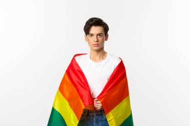Ciddi görünümlü cinsiyet değişkeni yüzü simli, lgbtq gökkuşağı bayrağı takan, beyaz arka planda duran biri.