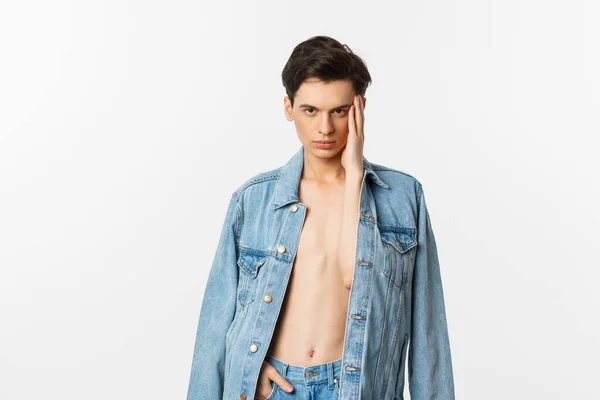 Όμορφος και αυθάδης γκέι άντρας που φοράει τζιν μπουφάν σε γυμνό κορμό, αγγίζει το πρόσωπό του και κοιτάζει με αυτοπεποίθηση την κάμερα, στέκεται πάνω από το λευκό φόντο — Φωτογραφία Αρχείου