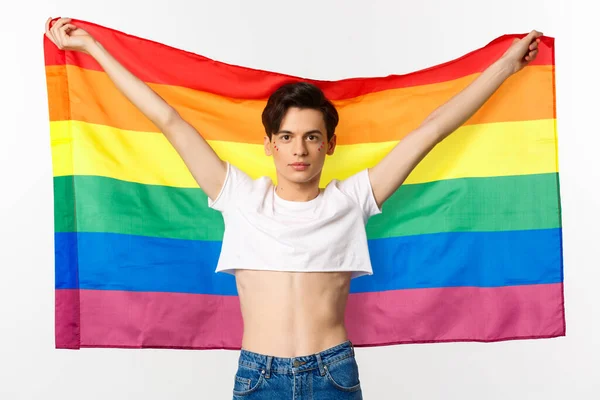 Fuera y orgulloso. feliz y confiado gay hombre con brillo en la cara levantando lgbtq arco iris bandera y sonriendo a cámara, de pie en la parte superior de la cosecha, fondo blanco —  Fotos de Stock