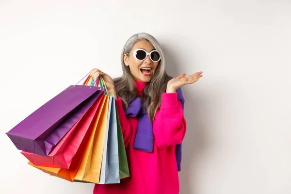 Stilvolle ältere asiatische Frau mit Sonnenbrille, die im Urlaub einkaufen geht, Papiertüten in der Hand und lächelnd vor weißem Hintergrund steht — Stockfoto