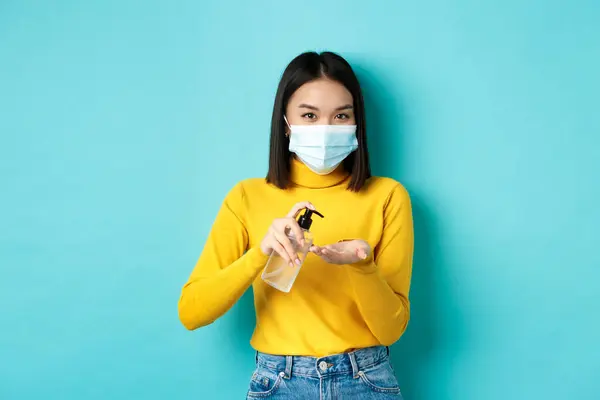 Covid-19, socialt avståndstagande och pandemikoncept. Le asiatisk kvinna i medicinsk mask med hjälp av förebyggande åtgärder från coronavirus, med hjälp av handdesinfektionsmedel, står över blå bakgrund — Stockfoto