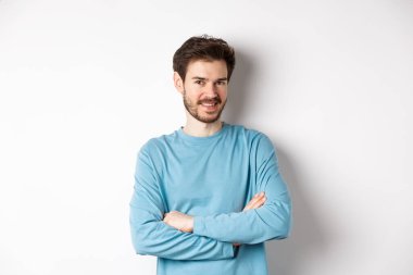 Sakallı, göğsünde çapraz kollar olan kendine güvenen genç bir adam ve kameraya gülümse, beyaz arka planda profesyonel gibi duruyor.