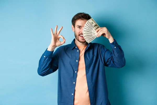 Joven alegre mostrando OK signo y cubrir la mitad de la cara con dólares, de pie sobre fondo azul — Foto de Stock