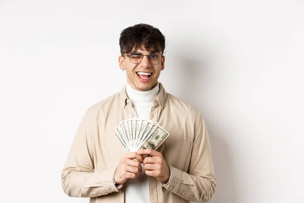 Guapo joven guiñando un ojo y mostrando billetes de dólar, sosteniendo fan de billetes de dinero y sonriendo complacido, haciendo dinero, de pie sobre fondo blanco — Foto de Stock