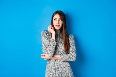 Uzun doğal saçlı, eski moda elbiseler giyen, düşünceli yüzüyle sol tarafa bakan, mavi arka planda duran düşünceli beyaz kız.