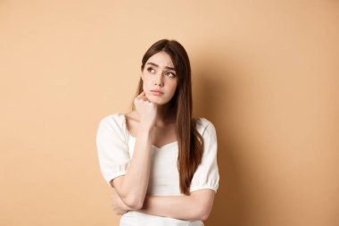 Üzgün genç bir kadın sol üst köşede dalgın görünüyor, üzgün ya da üzgün, bej arka planda tek başına duruyor.