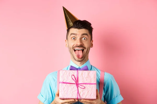 Holidays and celebration concept. Funny guy celebrating birthday, wearing party hat, holding b-day gift and showing tongue with happy face, pink background — Stock Photo, Image