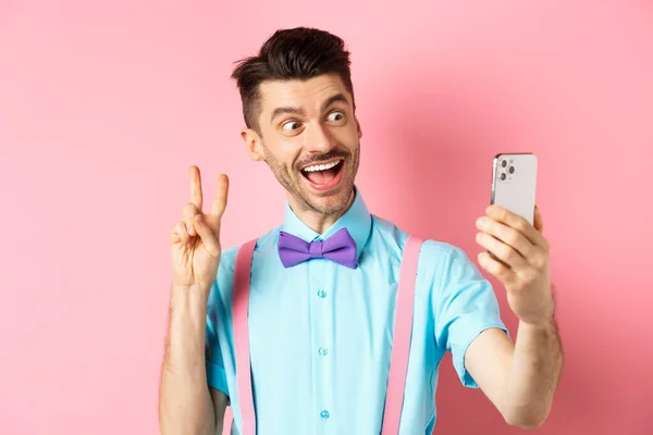 Technologický koncept. Vtipný muž s knírkem a motýlkem se selfie na smartphonu, ukazující mírové znamení a usmívající se na mobilní kameru, růžové pozadí — Stock fotografie