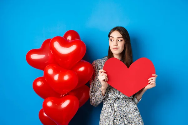 День святого Валентина. Романтична дівчина в одязі стоїть біля повітряних кульок і тримає велике червоне серце вирізати, мріяти про щось, стоячи на синьому фоні — стокове фото