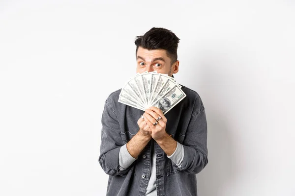 Emocionado tipo divertido escondiendo la cara detrás de billetes de dólar y sonriendo, mostrando dinero en efectivo, de pie sobre fondo blanco — Foto de Stock