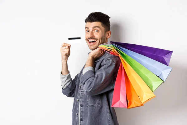 Chico alegre va de compras con tarjeta de crédito, sosteniendo bolsas en el hombro y sonriendo emocionado ante la cámara, fondo blanco — Foto de Stock