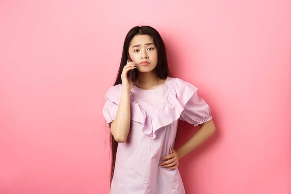 Sad asian girl calling someone, holding phone and talking, standing upset against pink background