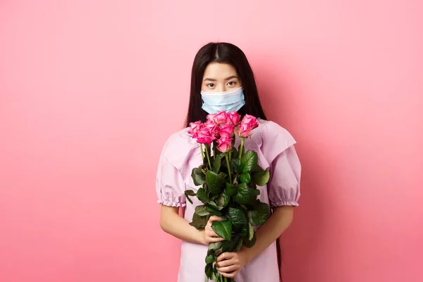 Young asian girl in medical mask holding flowers on Valentines day, receive bouquet of roses from lover, standing on pink background. Social distancing and covid-19 concept — Stock Photo, Image