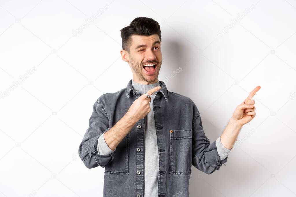 Handsome smiling guy making announcement, winking and pointing at upper right corner, showing logo, inviting to check out advertisement, standing on white background