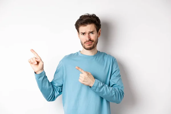 Jeune homme sceptique avec barbe grimaçant insatisfait, fronçant les sourcils et pointant vers le logo du coin supérieur gauche douteux, debout bouleversé sur fond blanc — Photo