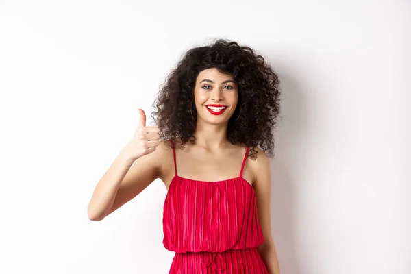 Vrolijke vrouw met rode lippen en elegante jurk, met duim omhoog en glimlachen, aanbevelen product, staande over witte achtergrond — Stockfoto