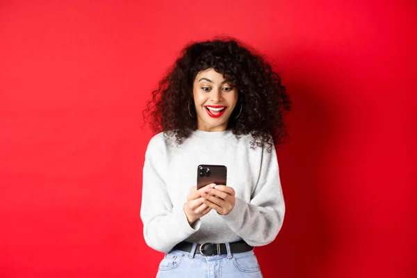Imagen de una mujer joven con el pelo rizado, leyendo el mensaje en el teléfono inteligente y sonriendo feliz, recibiendo buenas noticias en línea, de pie sobre fondo rojo — Foto de Stock
