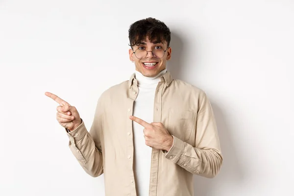 Homem bonito alegre em óculos sorrindo, apontando os dedos deixados no logotipo, mostrando propaganda com rosto animado, de pé sobre fundo branco — Fotografia de Stock