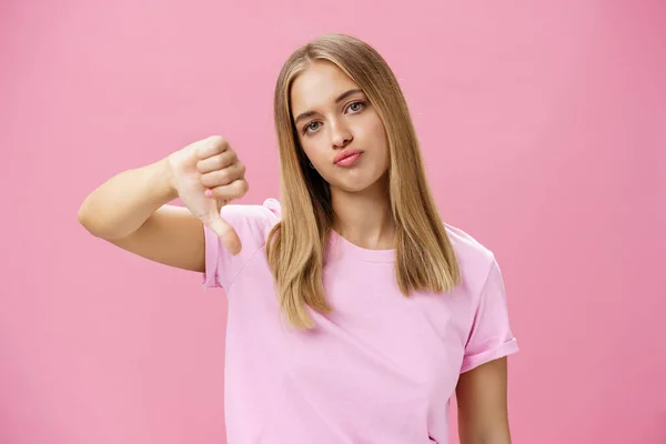 Displeased irritated and moody european woman with fair hair pursing lips annoyed showing thumbs down in dislike standing pissed and unimpressed giving negative feedback against pink wall — Stockfoto