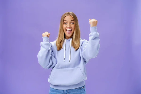 Allegro felice e solidale giovane ragazza con i capelli biondi e abbronzatura in caldo hoodide alzando pugni in allegria e trionfo sorridente dicendo sì celebrando obiettivo o successo su sfondo viola — Foto Stock