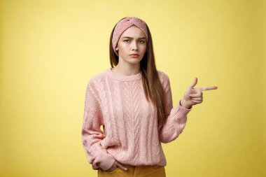 Pissed puzzled young attractive arrogant moody girlfriend in sweater, headband looking irritated, intense frowning pointing finger at copy space, directing person sits on her place, annoyed