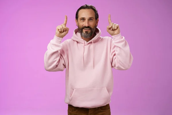 Schöne reife 50er Jahre Mann graues Haar Bart trägt stylische Hipster lose Kapuzenpullover zeigt Zeigefinger zeigt genial Ort hängen unwillig alt werden stehend positive Glück lila Hintergrund — Stockfoto