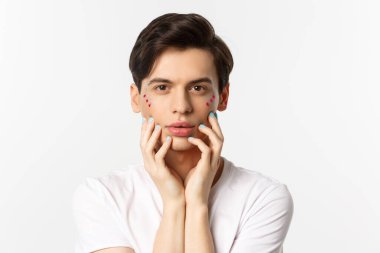 Millet, lgbtq ve güzellik konsepti. Yüzüne mavi ojeli parmaklarıyla dokunan, beyaz arka planda duran güzel eşcinsel adamın yakın çekimi.