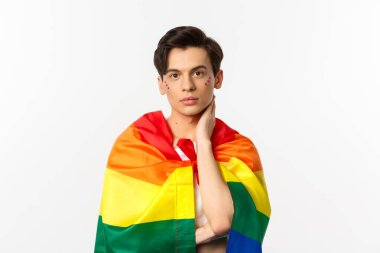 Yüzünde sim olan güzel bir gay adamın belden yukarı pozu, kendini gökkuşağı lgbt bayrağıyla sar ve yüzüne nazikçe dokun, kameraya bak, beyaz arkaplan