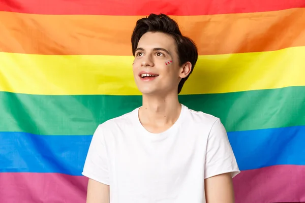 Tiro en la cintura de hombre gay feliz con brillo en la cara sonriendo, mirando soñador en la esquina superior izquierda, celebrando vacaciones de orgullo, luchando por la visibilidad, de pie contra la bandera del arco iris lgbtq —  Fotos de Stock