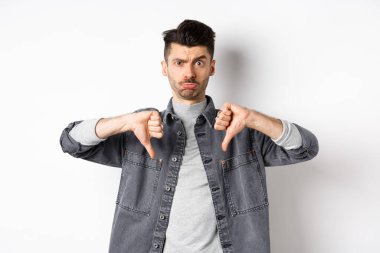 Hayal kırıklığına uğramış komik adam somurtuyor ve üzgün görünüyor, baş parmaklarını gösteriyor hoşnutsuz, beyaz arka planda duruyor.