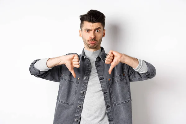 Teleurgesteld grappige man fronsen en kijken boos, duimen naar beneden ontevreden, staan op witte achtergrond — Stockfoto