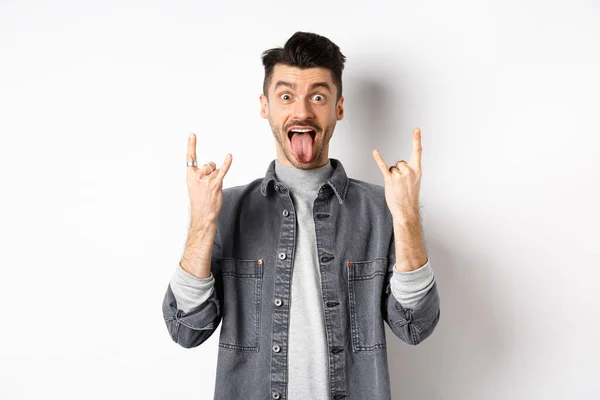 Excited funny guy showing tongue and heavy metal horns sign, enjoying party or event, having fun, standing on white background — Stock Photo, Image