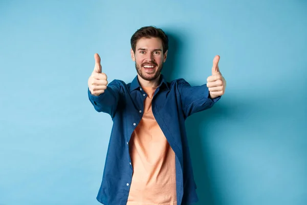 Joven guapo sonriendo y mostrando los pulgares hacia arriba en la aprobación, decir que sí con la cara feliz, de pie sobre fondo azul — Foto de Stock