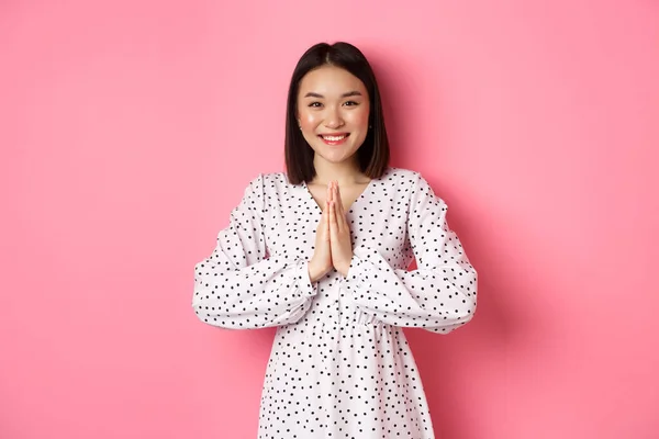 Bella donna asiatica che ti ringrazia, tenendosi per mano insieme nel gesto di apprezzamento, sorridendo felice alla macchina fotografica, in piedi grato su sfondo rosa — Foto Stock