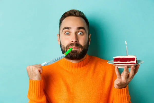 Close-up of funny adult man celebrating his birthday, holding bday cake with candle, blowing party wistle and rejoicing, standing over light blue background — Stock Photo, Image