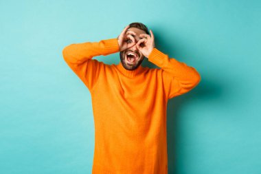 Komik ve mutlu bir adamın el ele tutuşup sahte gözlüklerden bakıp güldüğü turkuaz arka planda turuncu süveter giydiği görüntüsü.