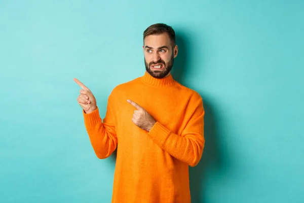 L'homme mécontent grince de quelque chose de mauvais, pointant et regardant quelque chose de dégoûtant ou d'étrange, l'espace de copie côté gauche, debout sur un fond turquoise — Photo