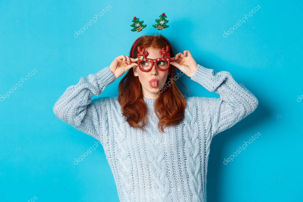 Christmas party and celebration concept. Silly redhead girl enjoying New Year, wearing funny glasses and headband, showing tongue and staring left at logo, blue background.