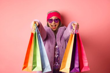 Mutlu Asyalı yetişkin kadın alışverişe çıkıyor, alışveriş torbalarıyla el ele tutuşuyor ve gülümsüyor, pembe arka planda dikiliyor.