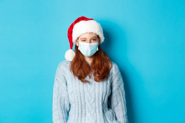 Concetto Natale, quarantena e Covid-19. Cheeky rossa modello femminile in maschera viso e cappello di Babbo Natale, strizzando l'occhio alla macchina fotografica, augurando Buon Natale, in piedi su sfondo blu — Foto Stock