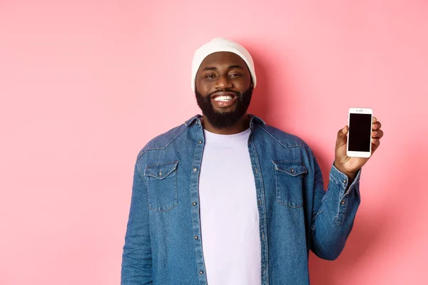 Chico hipster guapo en gorro y camisa de mezclilla sonriendo, mostrando la pantalla del teléfono móvil con la cara feliz, presentar la aplicación, de pie sobre el fondo rosa — Foto de Stock