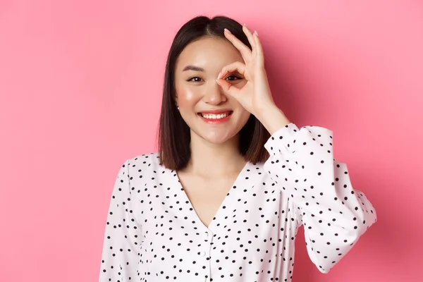 Kauneus ja elämäntapa käsite. Lähikuva söpö hymyilevä aasialainen tyttö osoittaa kunnossa merkki silmään, seisoo yli vaaleanpunainen tausta mekko — kuvapankkivalokuva